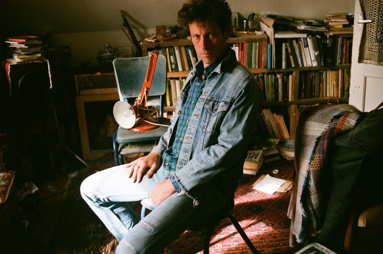 Sam Lee, wearing denim, sits in a cluttered room in front of a bookshelf